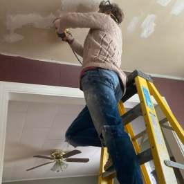 Person on ladder plastering ceiling with a tool in a room.