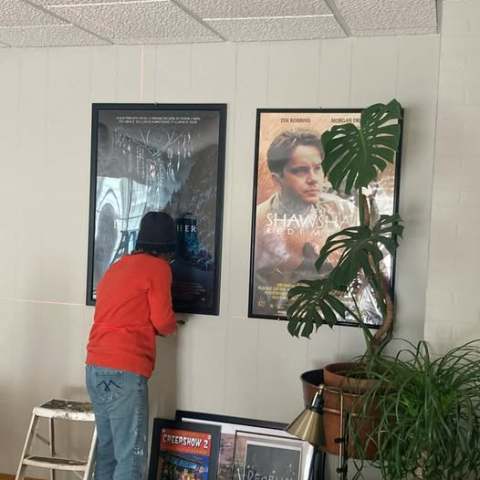 Person hanging a poster on the wall next to another movie poster, with a plant and furniture nearby.