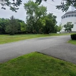 Paved road with grass and trees, water tower in background.