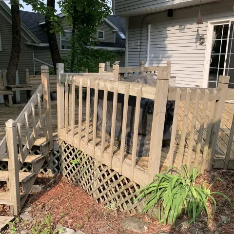 Wooden patio with grill and stairs, plants in foreground, house in background.