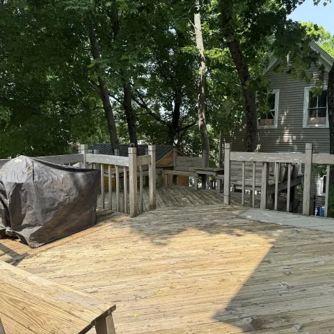 Wooden deck with a covered grill, trees, and a house in the background.