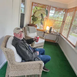Man sitting in wicker chair on a porch holding a drink can.
